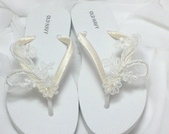 May include: A pair of white flip-flops with a satin strap and a white lace embellishment with pearl accents. The flip-flops are labeled "Old Navy".