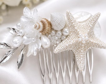 May include: A hair comb featuring a white starfish, small seashells, translucent white floral accents, silver-toned metal leaves, and clusters of white pearls. The decorative elements are arranged on a silver-coloured metal comb base.