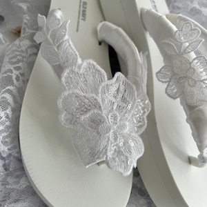 May include: A pair of white flip-flops with a white lace flower embellishment on each strap. The flip-flops are made by Old Navy.