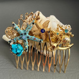 May include: Gold hair comb with a sea-themed design. It features a starfish, an anchor, a seashell, and coral, all adorned with blue and turquoise rhinestones.
