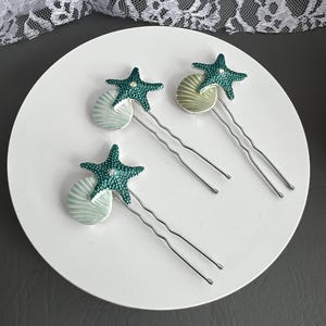 Puede incluir: Tres horquillas para el pelo con un diseño de temática marina. Cada horquilla presenta una estrella de mar turquesa con pequeños adornos y una concha de color verde claro. Las horquillas son plateadas y están dispuestas sobre una superficie circular blanca.