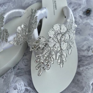May include: White flip flops with a silver floral embellishment and a white satin ribbon. The flip flops have the text "OLD NAVY" printed on the strap.