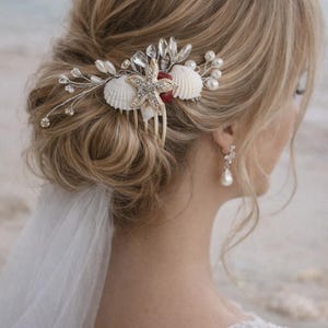 Op de afbeelding: Een bruidskam met een zeester, schelpen en kristallen. De kam is versierd met een sprankelende zeester, witte schelpen en heldere kristallen, perfect voor een bruiloft met een strandthema. De kam is geplaatst tegen blond haar.