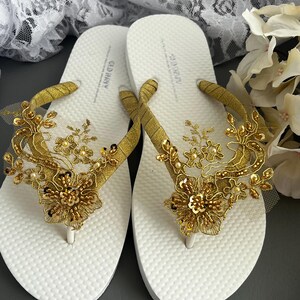 May include: A pair of white flip-flops decorated with gold lace and embellishments. The flip-flops are adorned with a floral design and have a textured sole.