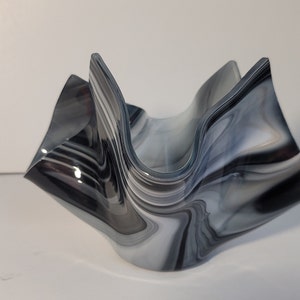 Puede incluir: Un cuenco decorativo de vidrio con un diseño ondulado único. El cuenco presenta un patrón veteado en tonos grises, negros y blancos. Los bordes del cuenco son curvos, creando una forma fluida y orgánica. El cuenco probablemente se utiliza con fines decorativos.