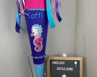 Pode incluir: Um cone escolar rosa e azul com um desenho de cavalo-marinho e o nome "Lotti" bordado. O cone é decorado com fitas e está em um suporte de metal. Uma placa diz "Endlich Schulkind Made by Deri".