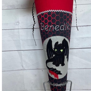 Könnte beinhalten: Ein rot-schwarzer Schultüte mit einem schwarzen Drachen-Design und dem Namen "Benedikt" bestickt. Die Tüte steht auf einem Metallständer.