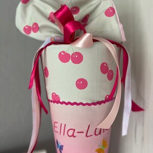Puede incluir: Un cono escolar rosa y verde con estampado de cerezas. El cono tiene una cinta rosa y una cinta blanca atadas alrededor. El cono tiene un borde festoneado rosa y el nombre "Ella-Lu" est&aacute; bordado en la parte delantera. El cono est&aacute; decorado con mariposas.