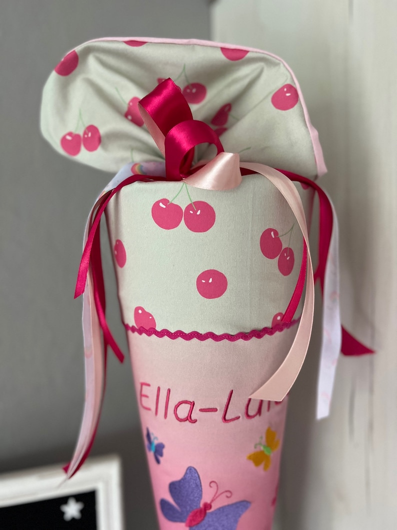 Puede incluir: Un cono escolar rosa y verde con estampado de cerezas. El cono tiene una cinta rosa y una cinta blanca atadas alrededor. El cono tiene un borde festoneado rosa y el nombre "Ella-Lu" est&aacute; bordado en la parte delantera. El cono est&aacute; decorado con mariposas.