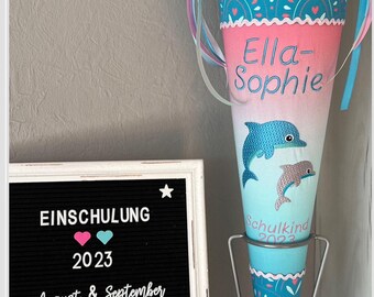 Puede incluir: Un cono escolar de color azul verdoso y rosa con un diseño de delfines. El cono tiene el nombre "Ella-Sophie" y el texto "Schulkind 2023" bordado. Un cartel dice "EINSCHULUNG 2023 August & September MADE BY DE RI beautiful".