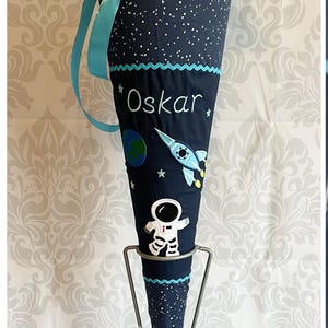 Könnte beinhalten: Ein blauer und weißer Schultüte mit Weltraum-Motiv. Die Tüte ist mit einer Rakete, einem Astronauten und der Erde bestickt. Die Tüte ist mit dem Namen "Oskar" personalisiert.