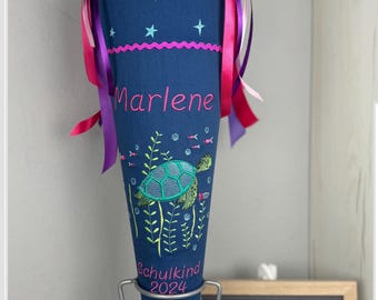 Puede incluir: Un cono escolar azul y rosa con un patrón de estrellas y un diseño de tortuga marina. El cono está decorado con cintas rosas y moradas y tiene el nombre "Marlene" y el texto "Schulkind 2024" bordado.