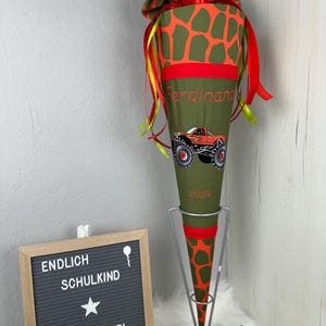 Könnte beinhalten: Eine grüne und orangefarbene Schultüte mit einem Monstertruck-Motiv und dem Namen "Ferdinand". Die Tüte ist mit rotem Band und einem Giraffenmuster verziert. Ein Schild zeigt "ENDLICH SCHULKIND" und "MADE BY DERI".