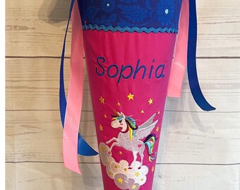 May include: A pink school cone with a blue and white unicorn design, the name "Sophia" embroidered on the front, and the year "2022" embroidered on the bottom. The cone is on a metal stand.