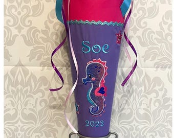 Pode incluir: Um cone escolar roxo e azul com um desenho de cavalo-marinho e o nome "Soe" bordado nele. O cone é decorado com glitter e lantejoulas e tem uma fita rosa amarrada ao redor. O cone está em um suporte de metal e o texto "MADE BY DERI" é visível na parte inferior da imagem.