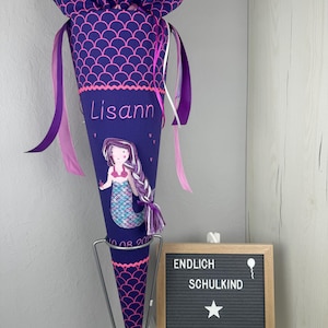 Könnte beinhalten: Ein lila-rosa Schultüte mit einem Meerjungfrauen-Motiv und dem Namen "Lisann" bestickt. Die Tüte ist mit rosa und weißen Bändern verziert und hat einen rosa Quasten. Die Tüte steht auf einem Metallständer. Ein Schild zeigt "ENDLICH SCHULKIND MADE BY DERI".