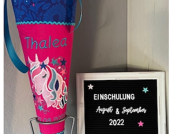 May include: A pink and blue school cone with a unicorn design and the name "Thalea" embroidered on it. The cone is on a metal stand and a black letter board with the text "EINSCHULUNG August & September 2022 VON MADE BY DERI" is in the background.