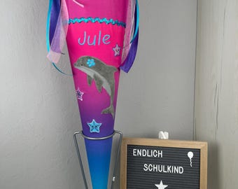 Puede incluir: Un cono escolar rosa y azul con un diseño de delfín y estrellas. El cono está decorado con el nombre "Jule" y el texto "ENDLICH SCHULKIND MADE BY DERI".