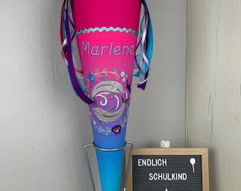 Puede incluir: Un cono escolar rosa y azul con un diseño de delfines y el nombre "Marlene" bordado en él. El cono está en un soporte de metal y tiene una cinta morada, azul y blanca atada a él. El cono está decorado con un ribete blanco y azul. Un letrero detrás del cono dice "Endlich Schulkind" y "Made by Deri".