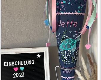 Puede incluir: Un cono escolar azul y rosa con un diseño de tortuga marina y el nombre "Jette" bordado. El cono está en un soporte y hay un letrero que dice "Einschulung 2023 August & September" con corazones y una estrella.