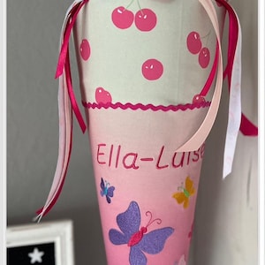 Puede incluir: Un cono escolar rosa y blanco con estampado de cerezas y el nombre "Ella-Luise" bordado. El cono est&aacute; decorado con mariposas de colores y est&aacute; sobre un soporte plateado.