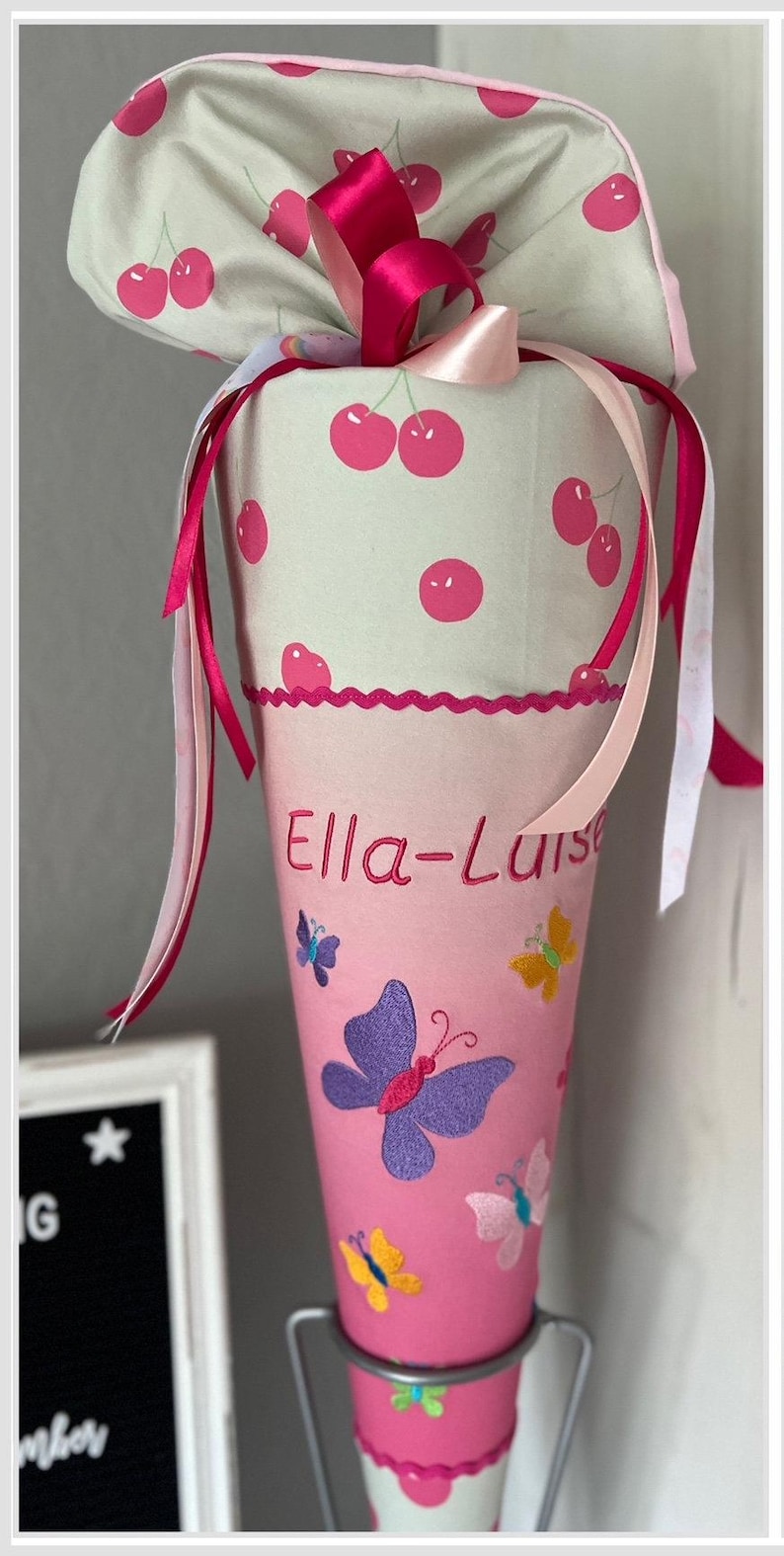 Puede incluir: Un cono escolar rosa y blanco con estampado de cerezas y el nombre "Ella-Luise" bordado. El cono est&aacute; decorado con mariposas de colores y est&aacute; sobre un soporte plateado.