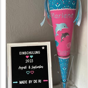 Könnte beinhalten: Ein rosa-blauer Schultüte mit einem Delfin-Design und dem Namen "Marlene" bestickt. Die Tüte steht auf einem Metallständer und hat ein Schild mit der Aufschrift "Einschulung 2023 August & September Made by de Ri beautiful".