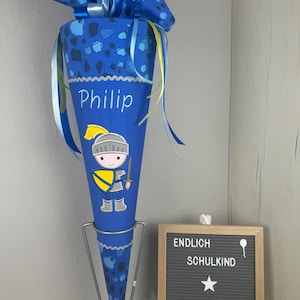 Könnte beinhalten: Ein blauer und gelber Schultüte mit einem Ritter-Motiv und dem Namen "Phillip" bestickt. Die Tüte ist mit einem blauen und gelben Band verziert und steht auf einem Metallständer. Im Hintergrund befindet sich eine schwarze Buchstaben-Tafel mit dem Text "ENDLICH SCHULKIND MADE BY DERI".