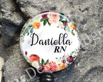 May include: A circular badge reel featuring a white background with a floral wreath of pink, orange, and dark red flowers and green leaves. The text "Daniella RN" is printed in black script and sans-serif font in the center of the badge.