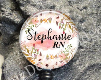 May include: A circular badge reel featuring a white background with a watercolor floral wreath in shades of pink, green, and brown. The text Stephanie RN is printed in black script and sans-serif font. It includes a black plastic housing and a clear strap.