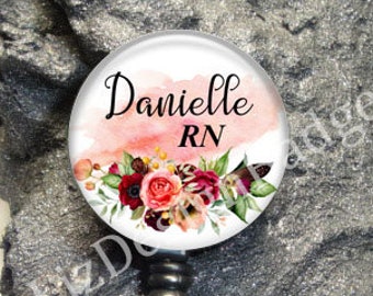 May include: A circular badge reel featuring a white background with a soft pink watercolor wash, a floral arrangement of red and dark pink roses, and the black text Danielle RN. The badge reel has a black plastic housing and a clear vinyl strap.