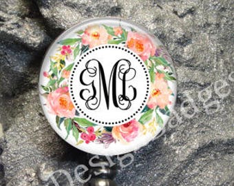 May include: A circular badge reel featuring a watercolor floral wreath design with pink and orange flowers and green leaves. In the center, a white circle with a black dotted border displays the black monogram letters ML in a script font.