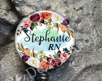 May include: A circular badge reel featuring a floral wreath design with red, white, and purple flowers and green leaves. The center displays the text Stephanie RN in black script and block lettering over a light blue watercolor background.