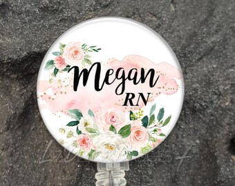 May include: A circular badge reel featuring a white background with a watercolor floral design in shades of pink, white, and green. The text "Megan" and "RN" is printed in black script and sans-serif font. It includes a clear plastic strap and metal snap.