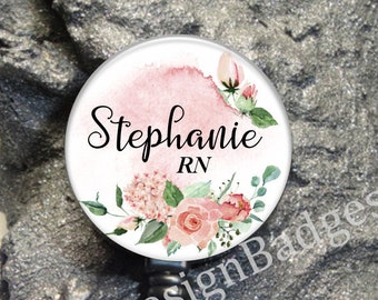 May include: A white round badge with a pink watercolor background and floral design. The text "Stephanie RN" is printed on the badge. The badge is attached to a black retractable badge reel.