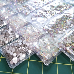 Könnte beinhalten: Sortiment aus klaren und schillernden Strasssteinen in verschiedenen Größen, verpackt in durchsichtigen Plastiktüten. Die Tüten sind in Reihen auf einer grünen Gitteroberfläche angeordnet.