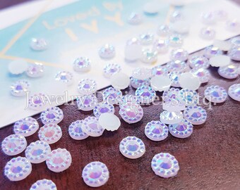 May include: A close-up of a group of iridescent white round rhinestones. The rhinestones have a clear center and a white outer edge. The rhinestones are scattered on a wooden surface.