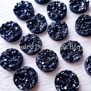 Könnte beinhalten: Eine Sammlung von 12 schwarzen runden Harz-Cabochons mit einer glitzernden, kristallartigen Textur. Die Cabochons haben einen Durchmesser von etwa 2,5 cm.