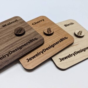 Op de afbeelding: Drie houten vierkanten met de woorden "Walnut", "Cherry" en "Birch" erop gegraveerd. Elk vierkant heeft een klein bladontwerp gegraveerd in het midden en de tekst "JewelryDesignersBtq." gegraveerd eronder.