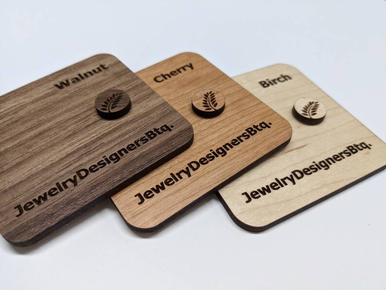 Op de afbeelding: Drie houten vierkanten met de woorden "Walnut", "Cherry" en "Birch" erop gegraveerd. Elk vierkant heeft een klein bladontwerp gegraveerd in het midden en de tekst "JewelryDesignersBtq." gegraveerd eronder.