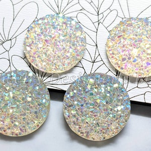 Può includere: Quattro cabochon rotondi iridescenti con una superficie scintillante simile al cristallo. I cabochon sono disposti su uno sfondo bianco con un motivo floreale. La superficie dei cabochon riflette più colori, creando un effetto cangiante.