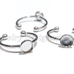 May include: Three silver-toned adjustable rings with circular bezel settings for cabochons. Each ring features a smooth, polished metal band with small spherical ends. The text Jewelry Designer's Btq. is overlaid across the image.