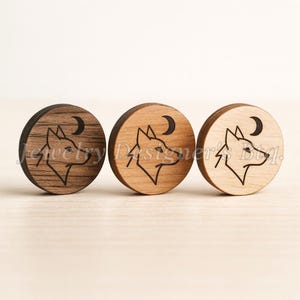 Puede incluir: Tres tapones redondos de madera con un diseño de lobo y luna creciente. Los tapones son de diferentes tonos marrones, con el lobo y la luna grabados en negro. Los tapones se muestran sobre una superficie de color claro.