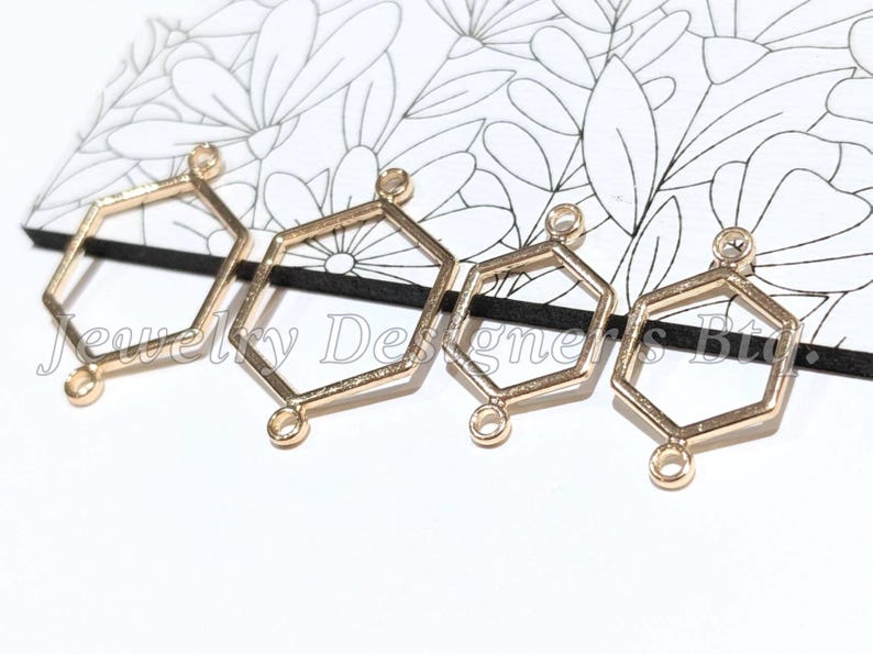 May include: Four gold-toned metal hexagon-shaped jewellery connectors arranged in a row. Each geometric frame features two small circular loops at opposite ends for attachment. The connectors vary slightly in size and have a smooth, metallic finish.