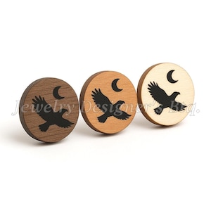 May include: Three round wooden earrings with a black eagle and crescent moon design. The earrings are in shades of brown and tan. The text "Jewelry Designers' Bdq." is visible.