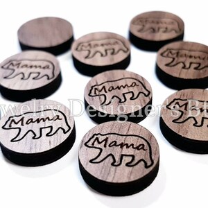 May include: Ten round wooden discs with a laser-etched design of a bear with the word "Mama" underneath. The discs are a light brown colour with a dark brown border.