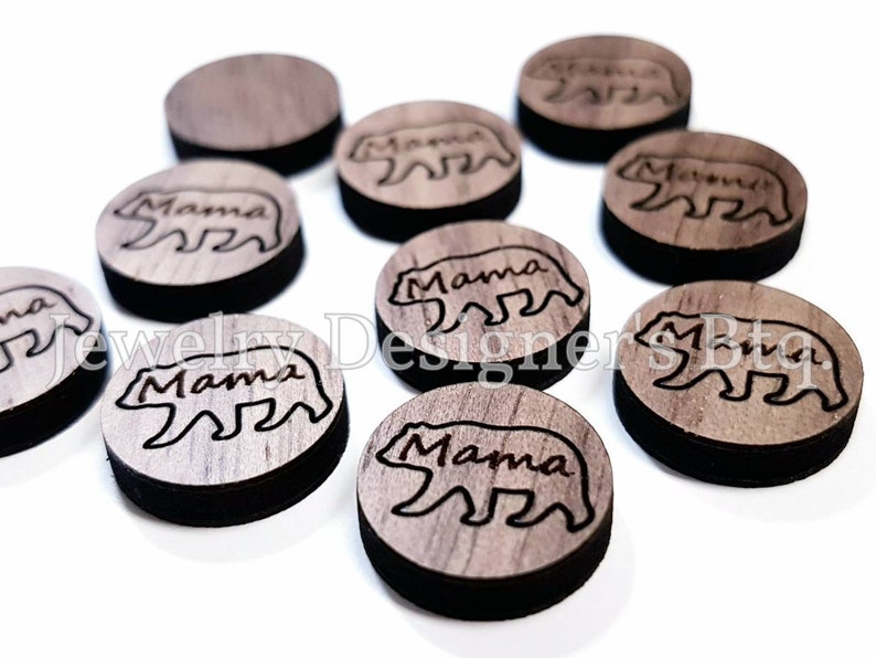 May include: Ten round wooden discs with a laser-etched design of a bear with the word "Mama" underneath. The discs are a light brown colour with a dark brown border.