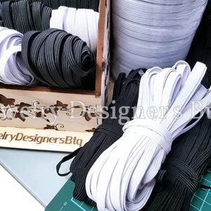 May include: Close-up of white and black elastic cord spools. The spools are arranged in a wooden box with a floral design and the text "@JewelryDesignersBtq".