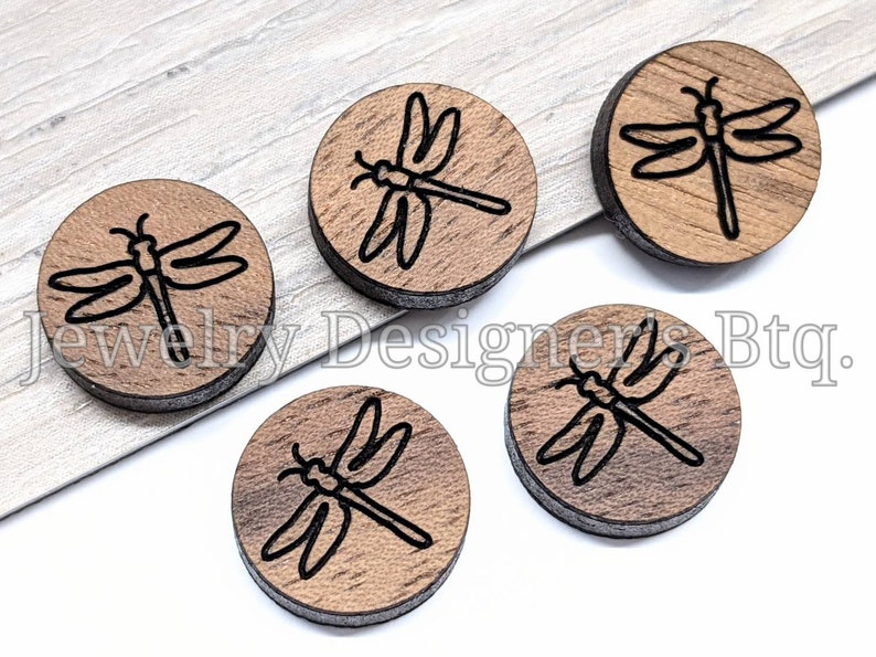 May include: Five wooden round ornaments with a black dragonfly design. The ornaments are approximately 1 inch in diameter.