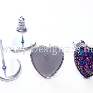 May include: Two heart-shaped earring settings, one with a purple and gold glitter design and the other empty. Both settings are made of silver metal.
